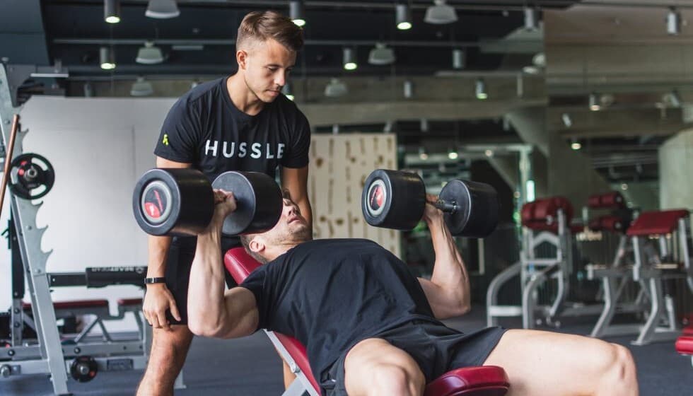Two members of staff wearing Hussle t-shirts sit next to each other in front of a boxing ring smiling at the camera.